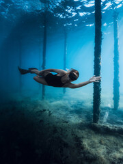 Freediver girl glides with fins dive under the pier in blue ocean. Female swims underwater dives between the pier pillars