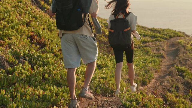 A man and a woman with backpacks enjoy a leisurely hike on a narrow trail amidst lush greenery on a sunny summer day, capturing the essence of adventure and tranquility.