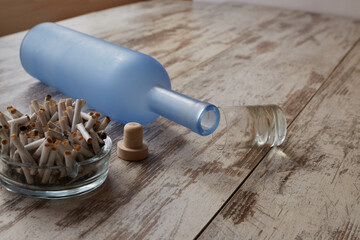 An overturned bottle of alcohol, an overturned glass, and an ashtray full of cigarette butts on the...