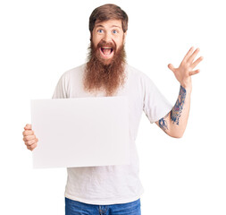 Handsome young red head man with long beard holding blank empty banner celebrating victory with happy smile and winner expression with raised hands