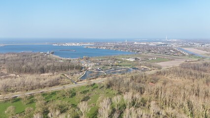 Aerial view of a coastal landscape with clear blue skies, featuring a calm bay, greenery, and infrastructure leading towards a distant industrial area.