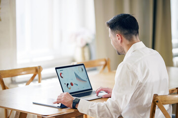 A meticulous man in a crisp white shirt reviews analytical data on his laptop, embodying professional diligence at a sunlit table.