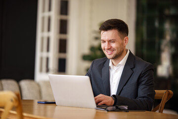 Fototapeta premium A businessman in a smart blazer and white shirt exudes a friendly demeanor while focusing on a laptop in a well-appointed office.