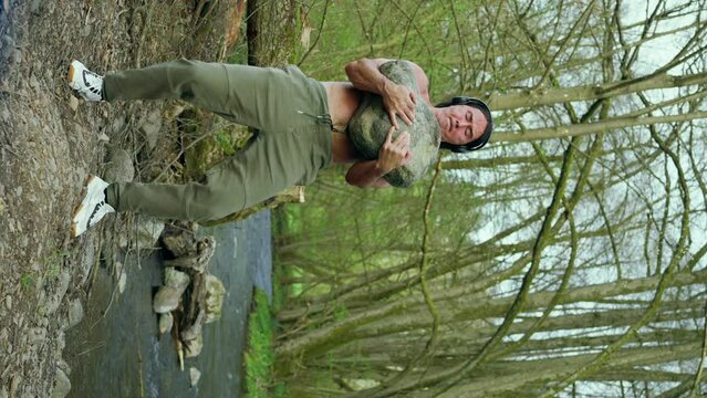 A man with a naked torso confidently near the river, lifts a large stone, slow motion, vertical view. A barechested man stands by water holding a large stone