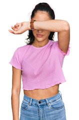 Hispanic teenager girl with dental braces wearing casual clothes covering eyes with arm, looking...