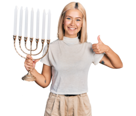 Beautiful young blonde woman holding menorah hanukkah jewish candle smiling happy and positive, thumb up doing excellent and approval sign