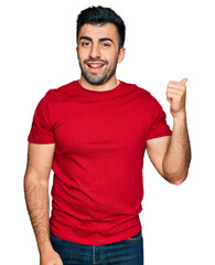 Hispanic man with beard wearing casual red t shirt smiling with happy face looking and pointing to the side with thumb up.