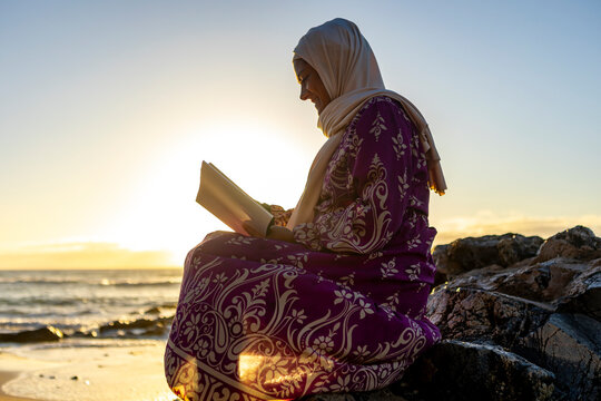 Serene Muslim woman reading a book on the beach at sunset