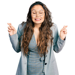 Young hispanic girl wearing business clothes and glasses gesturing finger crossed smiling with hope...