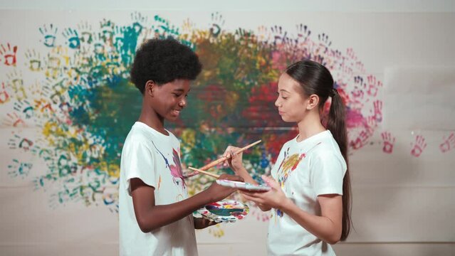 Smiling diverse children using paint brush painted color on each other white shirt shirt at colorful stained wall in art lesson. Represent exchanging experience, learning each other. Edification