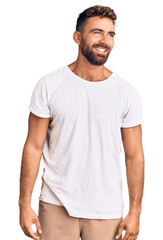 Young hispanic man wearing casual white tshirt looking away to side with smile on face, natural...