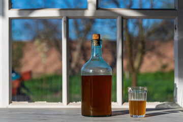 Big bottle with a drink made from fermented birch sap on the windowsill on a warm spring day, closeup. Traditional Ukrainian cold barley drink kvass in a glass jar and glass on table near yard - obrazy, fototapety, plakaty
