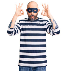 Young handsome man wearing burglar mask looking surprised and shocked doing ok approval symbol with...