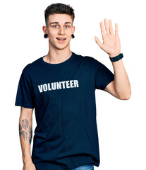 Young caucasian boy with ears dilation wearing volunteer t shirt waiving saying hello happy and smiling, friendly welcome gesture