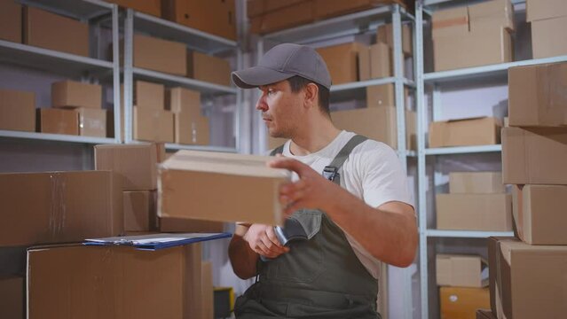 A male loader in uniform works with parcels using a barcode scanner in a warehouse with boxes