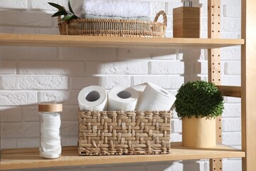Toilet paper rolls in wicker basket, floral decor and cotton pads on wooden shelf near white brick...