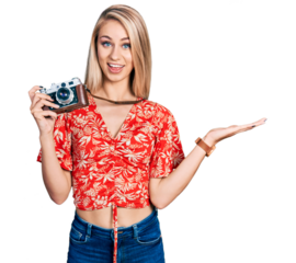 Beautiful young blonde woman holding vintage camera celebrating achievement with happy smile and winner expression with raised hand