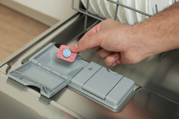 Man putting detergent tablet into open dishwasher indoors, closeup