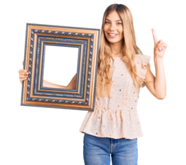 Beautiful caucasian woman with blonde hair holding empty frame surprised with an idea or question pointing finger with happy face, number one