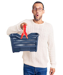 Young handsome man holding supermarket shopping basket scared and amazed with open mouth for...