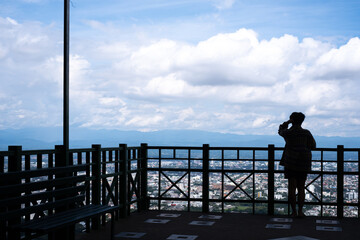 A woman is standing on a balcony overlooking a city. She is taking a picture of the city skyline with her cell phone