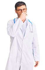 Fototapeta premium Handsome young man with bear wearing doctor uniform smelling something stinky and disgusting, intolerable smell, holding breath with fingers on nose. bad smell