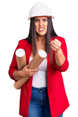 Young beautiful brunette woman wearing hardhat holding architect blueprint annoyed and frustrated shouting with anger, yelling crazy with anger and hand raised