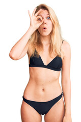 Young beautiful blonde woman wearing bikini doing ok gesture shocked with surprised face, eye looking through fingers. unbelieving expression.