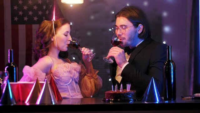 A man flirts with a beautiful woman at her birthday party, over a glass of red wine at the festive table, after the guests have left. They then share an exaggeratedly passionate kiss. Mid shot. 4k