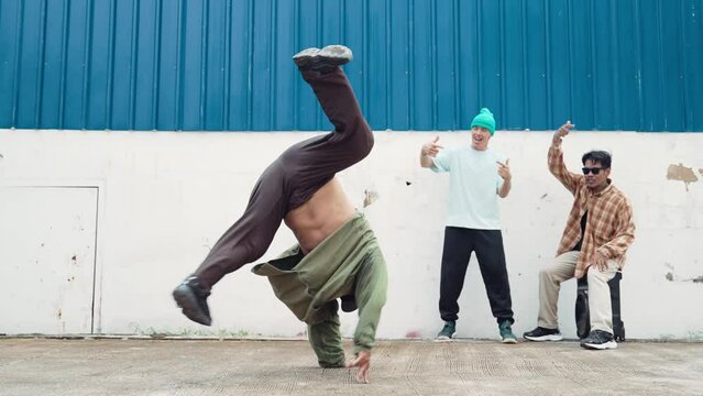 Skilled hipster doing cart wheel while multicultural friends encourage him. Hip-hop dancer moving foot step to music surrounded by group of hipster at wall near street. Outdoor sport 2024. hiphop.