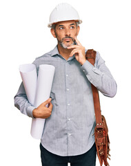 Middle age grey-haired man wearing safety helmet holding blueprints serious face thinking about...
