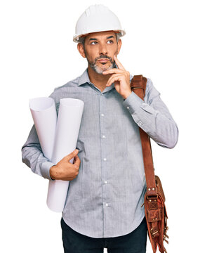 Middle age grey-haired man wearing safety helmet holding blueprints serious face thinking about question with hand on chin, thoughtful about confusing idea