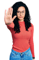 Young hispanic woman with curly hair wearing glasses doing stop sing with palm of the hand. warning...