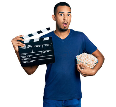 Young african american man eating popcorn holding film clapboard afraid and shocked with surprise and amazed expression, fear and excited face.