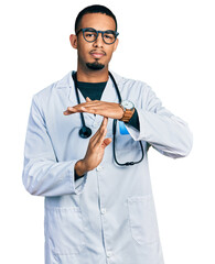 Young african american man wearing doctor uniform and stethoscope doing time out gesture with...