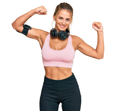 Young blonde woman wearing gym clothes and using headphones showing arms muscles smiling proud. fitness concept.