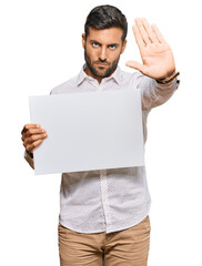 Handsome hispanic man holding blank empty banner with open hand doing stop sign with serious and...