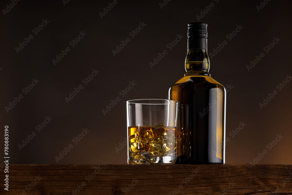 Sticker Whiskey with ice cubes in glass and bottle on wooden table, space for text