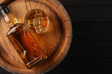 Whiskey with ice cubes in glass, bottle and barrel on black wooden table, top view. Space for text
