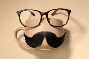 Artificial moustache, cup and glasses on beige background, top view