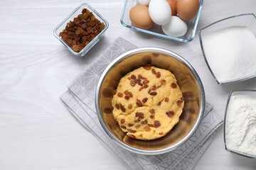 Dough with raisins in bowl and ingredients on white wooden table, flat lay