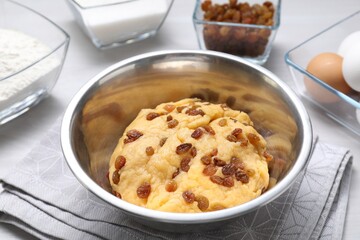 Dough with raisins in bowl and ingredients on white table, closeup