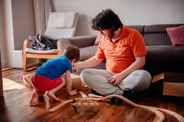 A child and an adult are absorbed in assembling a wooden toy train set on the floor, enjoying a...