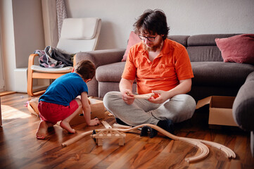 A child and an adult are absorbed in assembling a wooden toy train set on the floor, enjoying a playful and educational moment together