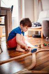 A focused young boy kneels on a sunny hardwood floor, guiding a blue toy train along its wooden track with keen interest and imagination