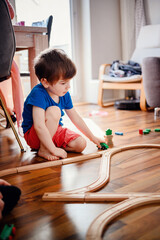 A focused young boy kneels on a sunny hardwood floor, guiding a blue toy train along its wooden track with keen interest and imagination