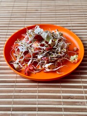 A fresh vegetable salad. Tomatoes, onions, apples, mound of sprouted mung beans in an orange bowl on bamboo background. Concept of diet, vegan, healthy products. Close up.