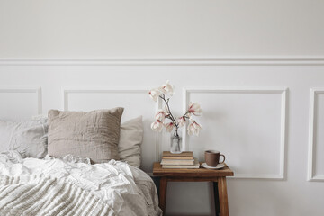 Elegant bedroom. Wooden night stand with fluted glass vase. Boolming magnolia tree branches. Cup of coffee on old books. Scandinavian interior. Linen bedding. White wall background, stucco decor. - obrazy, fototapety, plakaty