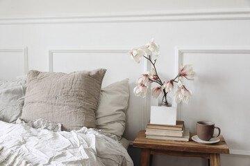 Blank greeting card, invitation mockup on old books. Elegant bedroom. Linen pillows, blanket. Wooden night stand, rippled glass vase. Blooming magnolia branches, cup of coffee. Scandinavian interior.