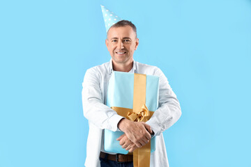 Mature man in party hat with birthday gift on blue background